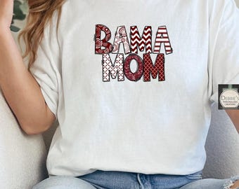 Puede incluir: Camiseta blanca con el texto "BAMA MOM" en un diseño decorativo y estampado. Las letras son en tonos rojos y blancos. La camiseta la lleva una persona.
