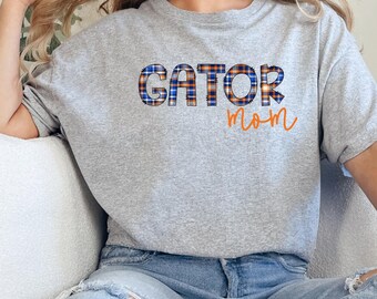 Puede incluir: Una camiseta gris jaspeado de manga corta con el texto GATOR en un patrón de cuadros azul y naranja, con la palabra mom escrita debajo en una fuente cursiva naranja. La camiseta se combina con unos vaqueros azul claro desgastados.