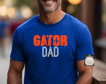 Puede incluir: Una camiseta de manga corta de color azul real con el texto GATOR DAD en letras de bloque naranjas y blancas. Un pequeño logotipo cuadrado con el texto DEBBIE'S PERSONALIZED CREATIONS es visible en el lado inferior derecho de la tela.