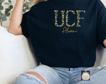 Puede incluir: Camiseta negra con la inscripción "UCF Alumni" en letras florales doradas. La camiseta es de cuello redondo y parece estar hecha de un material suave. El texto está en una fuente cursiva.