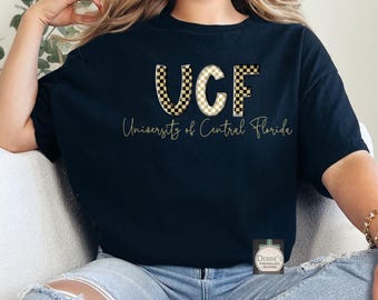 Puede incluir: Camiseta negra con las letras "UCF" en un diseño a cuadros negro y dorado. Debajo, el texto "University of Central Florida" en letras doradas. Una prenda casual para el día a día.