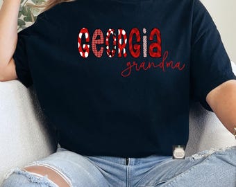 Puede incluir: Una camiseta negra de manga corta con la palabra GEORGIA en letras grandes, negritas y con patrones rojos y blancos, con la palabra grandma escrita debajo en una fuente roja cursiva más pequeña. La camiseta se combina con jeans azul claro.