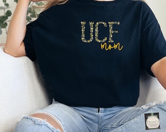 Puede incluir: Una camiseta negra de manga corta con el texto UCF en un patrón floral amarillo y negro, con la palabra mom escrita en cursiva amarilla debajo. La camiseta se combina con unos vaqueros de mezclilla azul claro desgastados con rodillas rotas.