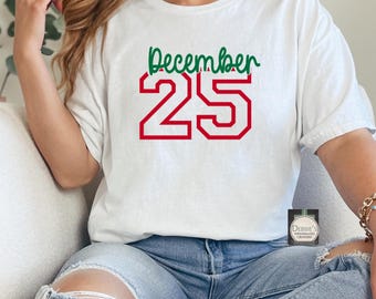 Puede incluir: Una camiseta blanca de manga corta con la palabra December en cursiva verde sobre el número 25 en letras de bloque rojas grandes con un contorno blanco. La camiseta tiene cuello redondo y un ajuste relajado.