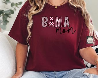 Puede incluir: Camiseta burdeos con la inscripción "BAMA mom". La palabra "BAMA" está en negrita, con un patrón de cuadros, mientras que "mom" está en cursiva. Esta camiseta informal es adecuada para el uso diario. La camiseta es de color rojo oscuro.