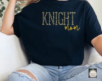 Puede incluir: Camiseta negra con el texto "KNIGHT mom" en letras doradas de purpurina. La camiseta la lleva puesta una persona sentada.