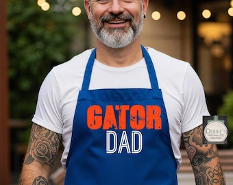 Puede incluir: Un delantal azul con las palabras "GATOR DAD" en naranja y blanco. El delantal está atado a la cintura y lo lleva una persona que viste una camiseta blanca. La persona tiene tatuajes en los brazos.