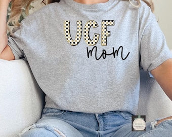 Puede incluir: Una camiseta gris de manga corta con el texto "UCF mom" en escritura negra y un estampado a cuadros. Las letras "UCF" tienen un estampado a cuadros negro y beige. La camiseta está hecha de un material suave.