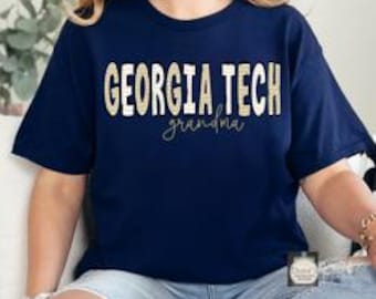 Puede incluir: Una camiseta azul marino de manga corta con el texto GEORGIA TECH en una fuente texturizada dorada y blanca, con la palabra grandma escrita en cursiva dorada debajo. La camiseta se combina con unos vaqueros azul claro desgastados.