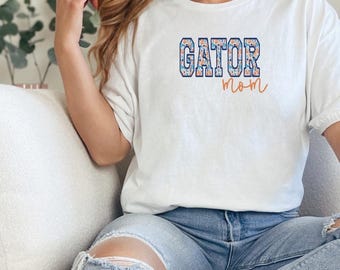 Puede incluir: Una camiseta blanca de manga corta con la palabra GATOR en una fuente de bloque floral azul y naranja, con la palabra mom escrita debajo en una letra cursiva naranja. La camiseta se combina con unos vaqueros azul claro desgastados.