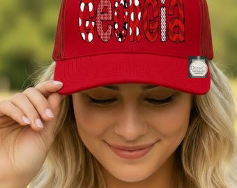 Puede incluir: Una gorra de estilo camionero de color rojo brillante con parte trasera de malla y panel frontal sólido. La palabra GEORGIA está impresa en el frente en letras grandes y estilizadas, cada una con un patrón único en rojo, blanco y negro.