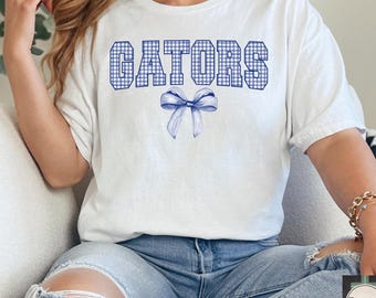 Puede incluir: Camiseta blanca con un estampado de cuadros azules y blancos que deletrea "GATORS" con un lazo azul de cuadros debajo.