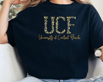 Puede incluir: Una camiseta negra de manga corta con las letras UCF en un patrón floral amarillo y blanco. Debajo de las letras, el texto University of Central Florida está impreso en una fuente cursiva amarilla. La camiseta tiene cuello redondo y corte holgado.