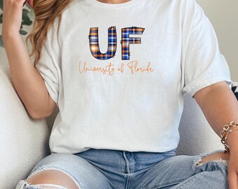Puede incluir: Una camiseta blanca de manga corta con las letras UF en un patrón de cuadros azul y naranja, con el texto University of Florida escrito en cursiva naranja debajo de las letras. La camiseta se combina con unos vaqueros azul claro desgastados.