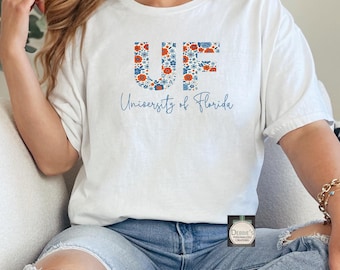 Puede incluir: Una camiseta blanca de manga corta con las letras UF rellenas de un estampado floral azul y naranja. Debajo de las letras, el texto University of Florida está impreso en una fuente cursiva azul. La camiseta se combina con unos vaqueros azul claro.