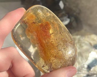May include: A clear, amber-colored stone with an embedded reddish-brown inclusion. The stone is held in a hand, showcasing its translucent quality and the intricate details within. The stone's shape is rectangular with rounded edges.