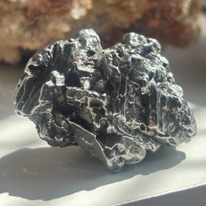Pu&ograve; includere: Un oggetto metallico scuro, di forma irregolare, con una superficie ruvida e strutturata. L'oggetto ha un colore argento e sembra essere una formazione naturale, forse un minerale o una meteorite. L'immagine &egrave; ben illuminata.