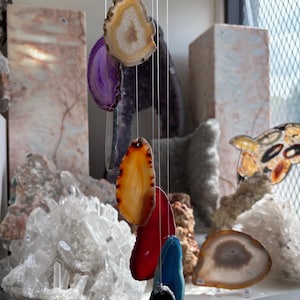 May include: A colorful agate wind chime with slices of purple, orange, red, blue, and black stones. The agate slices are suspended by thin strings. The background features various crystals and stone formations on a shelf.