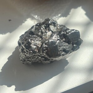 Pu&ograve; includere: Un oggetto metallico scuro di forma irregolare con una superficie ruvida e strutturata. L'oggetto &egrave; di colore grigio scuro e sembra essere una roccia o un minerale. L'oggetto proietta un'ombra su una superficie bianca.