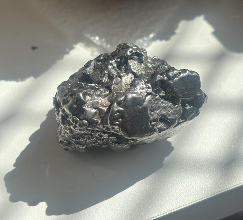 Pu&ograve; includere: Un oggetto metallico scuro di forma irregolare con una superficie ruvida e strutturata. L'oggetto &egrave; di colore grigio scuro e sembra essere una roccia o un minerale. L'oggetto proietta un'ombra su una superficie bianca.