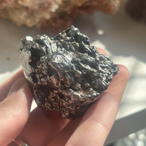 Pu&ograve; includere: Una roccia metallica di forma irregolare con una superficie strutturata. La roccia &egrave; di colore grigio argento scuro e sembra essere un campione minerale. L'oggetto &egrave; tenuto in una mano.
