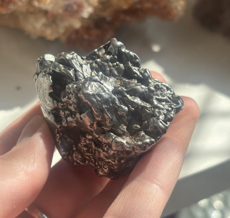 Pu&ograve; includere: Una roccia metallica di forma irregolare con una superficie strutturata. La roccia &egrave; di colore grigio argento scuro e sembra essere un campione minerale. L'oggetto &egrave; tenuto in una mano.