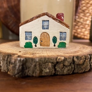 May include: A miniature ceramic house with a brown roof and chimney, blue windows, and a wooden door. The house sits on a rustic, round wooden slice, showcasing a charming, handcrafted aesthetic.
