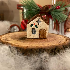 May include: A miniature ceramic Christmas house with a brown roof, door, and window. The house is decorated with a star, holly, and a red chimney. It sits on a wooden slice, surrounded by white fluff, with a festive background.