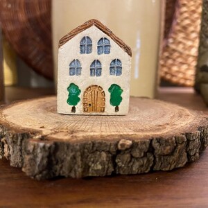 May include: A miniature ceramic house with a brown roof, blue windows, and a wooden door. Two green trees flank the door. The house sits on a rustic, round wooden slice, showcasing a charming, handcrafted aesthetic.