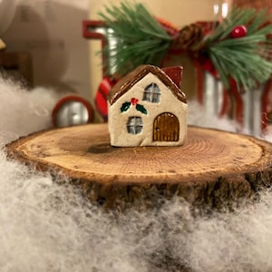 May include: A miniature ceramic house with a brown roof, a wooden door, and two windows. The house is decorated with holly and berries and sits on a wooden slice. The scene is set against a backdrop of white fluff and Christmas decorations.