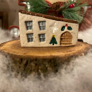 May include: A small, ceramic house figurine with a cream-colored exterior, brown roof, and decorative windows. The house features a green Christmas tree, a wooden door, and holly accents, all set on a wooden slice. The scene evokes a festive holiday theme.