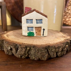 May include: A miniature ceramic house with a red chimney, blue windows, and a brown door. The house is set on a rustic wooden slice, with a small green bush and a black lamppost. The house is a charming decorative item.