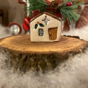 May include: A small, ceramic house figurine with a brown roof, door, and chimney. The off-white house features a star, holly, and a window. It sits on a wooden slice, surrounded by faux snow, suggesting a holiday theme.