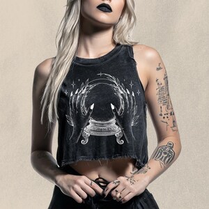 May include: Black crop top with a distressed hem featuring a white graphic of a crystal ball held by two hands. The design includes stars and a decorative base. The top is worn by a person with tattoos.
