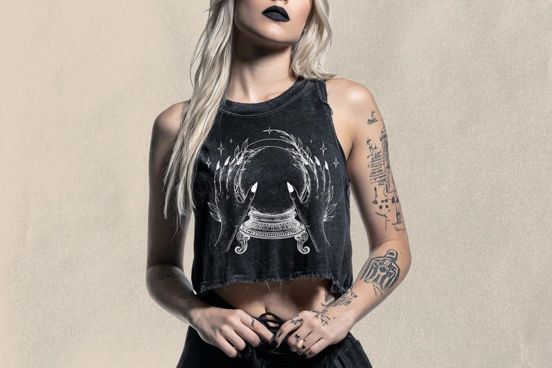 May include: Black crop top with a distressed hem featuring a white graphic of a crystal ball held by two hands. The design includes stars and a decorative base. The top is worn by a person with tattoos.