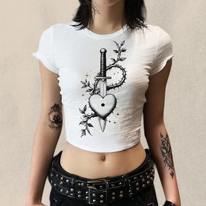 May include: White crop top with a black and white graphic of a heart pierced by a sword, surrounded by thorns and leaves. The top has short sleeves and a round neckline. The model is wearing a black studded belt.