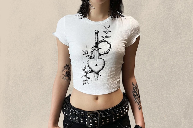 May include: White crop top with a black and white graphic of a heart pierced by a sword, surrounded by thorns and leaves. The top has short sleeves and a round neckline. The model is wearing a black studded belt.