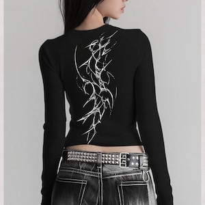 May include: Black long-sleeve top with a white abstract design on the back. The top is ribbed and form-fitting. The model is wearing a studded belt and dark wash jeans. The image also includes the text "high res" and "300 DPI".
