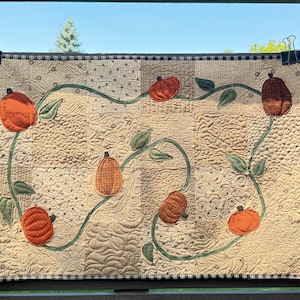 May include: A rectangular quilted wall hanging featuring a beige patchwork background with various stitched textures. Six fabric pumpkins in shades of orange and brown are connected by a winding green vine with leaves, finished with a black and white check border.