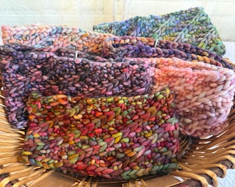 Puede incluir: Una colección de cinco diademas gruesas tejidas a mano con lana gruesa en varios colores variados, incluyendo tonos de rosa, morado, verde, naranja y azul, todas exhibidas juntas dentro de una cesta de mimbre tejida.
