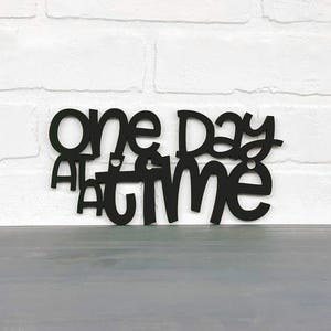 May include: Black wooden sign with the text "one day at a time" in a whimsical font.