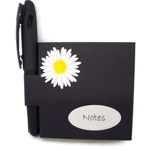 May include: Black notepad with a white daisy and the word "Notes" in a white oval. A black pen is attached to the notepad.