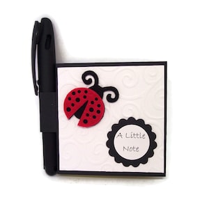 May include: A black notepad with a black pen attached to the side. The notepad has a white background with a black border and a black and white swirl pattern. A red ladybug with black spots is on the front of the notepad. The notepad has a black circle with the text "A Little Note" in white.