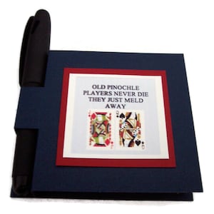 May include: A blue notepad with a red border and a white inner border. The notepad has a pen holder and the text "Old Pinochle Players Never Die They Just Meld Away" with two playing cards.