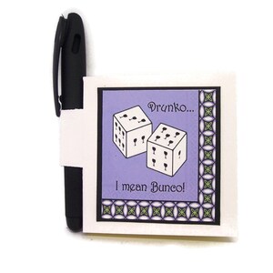 May include: A black pen with a white paper holder featuring a purple square with dice illustrations and the text "Drunko... I mean Bunco!". The dice have wine glass pips. The paper holder is decorated with a green and black geometric border.