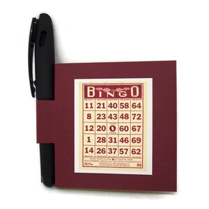 May include: A burgundy card with a white bingo card printed on it. The bingo card has the numbers 11, 21, 40, 58, 64, 8, 23, 43, 48, 72, 12, 20, 0, 60, 67, 1, 28, 31, 46, 69, 14, 26, 37, 57, 62. The card also has the text "BINGO" printed on it.
