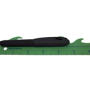 May include: A green plastic ruler with a black pen on top. The ruler has a wave-shaped edge and is marked with numbers from 1 to 6. The text "MADE IN THE USA" is printed on the ruler.