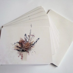Puede incluir: Una pila de tarjetas blancas en blanco con una sola tarjeta que presenta una ilustraci&oacute;n de acuarela de un ramo de flores secas en tonos de marr&oacute;n, beige y blanco.