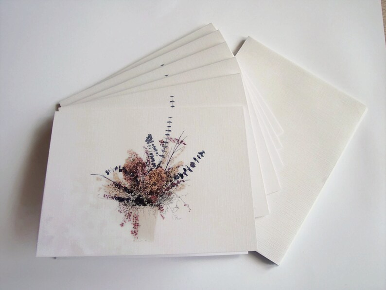 Puede incluir: Una pila de tarjetas blancas en blanco con una sola tarjeta que presenta una ilustraci&oacute;n de acuarela de un ramo de flores secas en tonos de marr&oacute;n, beige y blanco.