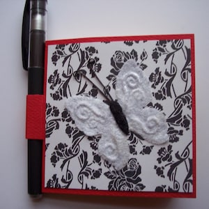 May include: A red and black notepad with a white butterfly design. The butterfly has black accents and is on a black and white damask pattern background.